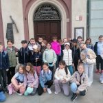 Grupa dzieci pozuje i uśmiecha się przed zabytkowym budynkiem z łukowatymi oknami i drewnianymi drzwiami.