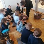 Grupa dzieci siedzi na podłodze, a dorosły pokazuje im instrument muzyczny w muzeum.