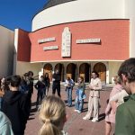 Grupa ludzi stoi w kręgu przed okrągłym budynkiem z łukowatymi, drewnianymi drzwiami i symbolami religijnymi.