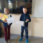 Dwoje dzieci stoi w pomieszczeniu, uśmiecha się, trzyma certyfikaty i nagrody w rękach, w otoczeniu klasy.