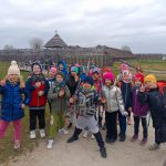 Grupa uśmiechniętych dzieci w kolorowych ubraniach pozuje na zewnątrz drewnianej fortecy w pochmurny dzień.