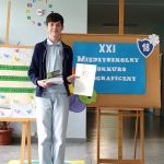 Uśmiechnięty nastolatek trzymający certyfikat, stojący przy znaku konkursu ortograficznego w języku polskim.