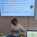 Chłopiec układa kostkę Rubika przy stole; na ekranie za nim widoczne są jego najlepsze czasy w różnych konkurencjach z kostką.