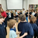 Grupa dzieci i nauczyciel wchodzą ze sobą w interakcję i rozmawiają w klasie pełnej regałów z książkami.