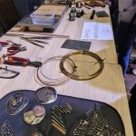 Na stole wystawiono drut metalowy, szczypce, ręcznie robioną biżuterię z metalu i szyld z napisem „Ręczna obróbka metalu”.