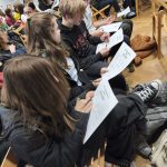 Grupa studentów siedząca na krzesłach i wypełniająca dokumenty podczas wydarzenia lub warsztatów.