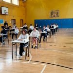 Uczniowie siedzą w indywidualnych ławkach na sali gimnastycznej i pod nadzorem piszą egzamin.