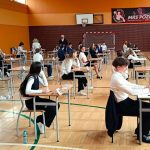 Uczniowie siedzą w ławkach w sali gimnastycznej i piszą egzamin. Nauczyciele stoją w pobliżu i obserwują klasę.