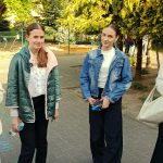 Cztery nastolatki stoją na zewnątrz, na wybrukowanej ścieżce, rozmawiają i uśmiechają się, w tle widać drzewa i plac zabaw.