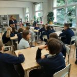 Nauczyciel stoi z przodu klasy i przemawia do uczniów siedzących w grupach w ławkach.