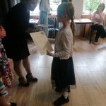 Kobieta wręcza certyfikat młodej dziewczynce w klasie, a wokół niej kręcą się inni ludzie.