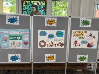 Trzy plakaty projektów uczniowskich na szarych tablicach, każdy z kolorowymi rysunkami i polskim tekstem, wyeksponowane w pomieszczeniu.