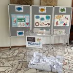 Przezroczyste pudełko wypełnione karteczkami stoi przed kolorowymi plakatami i szarymi tablicami w wyłożonym kafelkami pomieszczeniu.