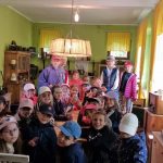 Grupa dzieci w kapeluszach siedzi i stoi w jasnej, zabytkowej klasie z zielonymi ścianami i drewnianymi meblami.