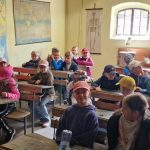 Dzieci siedzą przy starych drewnianych ławkach w klasie, w której na ścianach wiszą mapy świata, a przez okno wpada światło słoneczne.