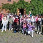 Grupa dzieci i dwójka dorosłych pozuje i uśmiecha się na zewnątrz ceglanego budynku porośniętego zielonym bluszczem.