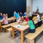 Dzieci siedzące przy drewnianych stołach wykonują prace plastyczne lub zadania w klasie pod okiem osoby dorosłej.