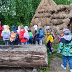 Grupa dzieci z plecakami zbiera się przy strzechach i dużych kłodach drewna na zewnątrz.