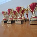 Siedem złotych i czerwonych trofeów ustawiono na drewnianym stole, na tle ściany i ekranu.