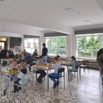 Dzieci w jasnym pokoju grają w szachy przy stołach, z dużymi oknami i pod nadzorem dorosłych.