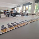 Dzieci grają w szachy w jasnym pokoju, na stole na pierwszym planie znajdują się certyfikaty i figury szachowe.