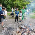 Dzieci i dorośli stoją wokół ogniska w lesie, piekąc jedzenie na patykach.