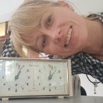 Uśmiechnięta kobieta w kraciastej koszuli pozuje obok zabytkowego analogowego zegara szachowego na stole.