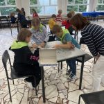 Dzieci wspólnie układają puzzle przy stole, a dorosły obserwuje ich pracę w jasnym, przestronnym pokoju.