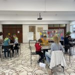 Dzieci siedzą w małych grupach przy stołach w jasnym pomieszczeniu i pod nadzorem dorosłych wykonują zadania.