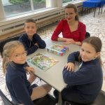 Czwórka dzieci i kobieta siedzą przy stole i grają w grę planszową w jasnym pomieszczeniu z dużymi oknami.