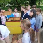 Grupa dzieci zbiera się wokół żółtego ula ustawionego na zewnątrz i obserwuje dorosłego trzymającego ramkę w kształcie plastra miodu.