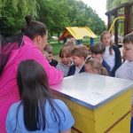 Grupa dzieci słucha kobiety ubranej na różowo, stojącej przy żółtym pudełku na placu zabaw.