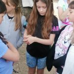 Grupa dzieci stoi na zewnątrz; jedna dziewczynka o długich brązowych włosach patrzy w dół, trzymając się za ręce.