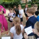 Grupa dzieci zbiera się na zewnątrz, trzyma w rękach gazety i słucha dorosłego w różowym stroju.