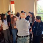 Grupa studentów gromadzi się wokół starszego mężczyzny w pomieszczeniu, słuchając go uważnie w pobliżu okna.