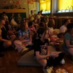 Dzieci siedzą blisko siebie na podłodze i poduszkach, oglądając film w słabo oświetlonej klasie.