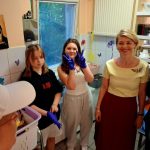 Cztery osoby stoją w kuchni, dwie dziewczyny w niebieskich rękawiczkach, jedna uśmiechnięta i pokazująca znak pokoju.