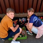 Dwoje dzieci przykuca przy skrzynkach z różnymi owocami i warzywami na ziemi, uśmiechając się do kamery.