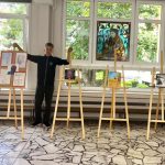 Chłopiec stoi w pomieszczeniu obok kilku drewnianych sztalug prezentujących różne dzieła sztuki.
