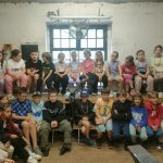 Grupa dzieci siedzi na ławkach w rustykalnym pokoju, twarzą do kamery, z oknem i wentylatorem za nimi.