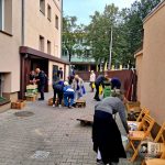 Ludzie sortujący i pakujący pudełka z żywnością na zewnątrz budynku, z drzewami i szkołą w tle.