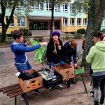 Trzy osoby sortują ubrania i przedmioty w pudełkach na ławce na zewnątrz, w pobliżu budynku szkoły.