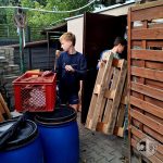 Dwóch chłopców przenosi drewnianą paletę i skrzynię obok niebieskich beczek i drewnianego płotu na zewnątrz.