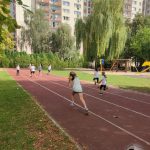 Dzieci biegające na bieżni w parku z budynkami mieszkalnymi i drzewami w tle.