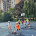 Dzieci grają w piłkę nożną na boisku zewnętrznym z budynkami mieszkalnymi i drzewami w tle.