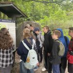 Grupa uczniów czyta znak informacyjny w zalesionym miejscu na zewnątrz.