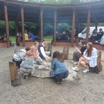 Grupa nastolatków siedzi wokół ogniska, piekąc jedzenie, a inni zgromadzili się pod drewnianą wiatą.