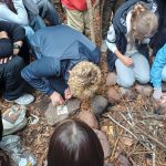 Dzieci zebrały się w kręgu na zewnątrz, obserwując, jak chłopiec używa krzesiwa do rozpalenia ognia na kamieniu.
