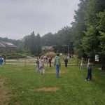 Grupa ludzi stoi i spaceruje po trawiastym terenie w pobliżu drzew i budynków pod zachmurzonym niebem.