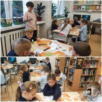 Dzieci siedzą przy stolikach w klasie, pracując z książkami i papierami, pod nadzorem nauczyciela.